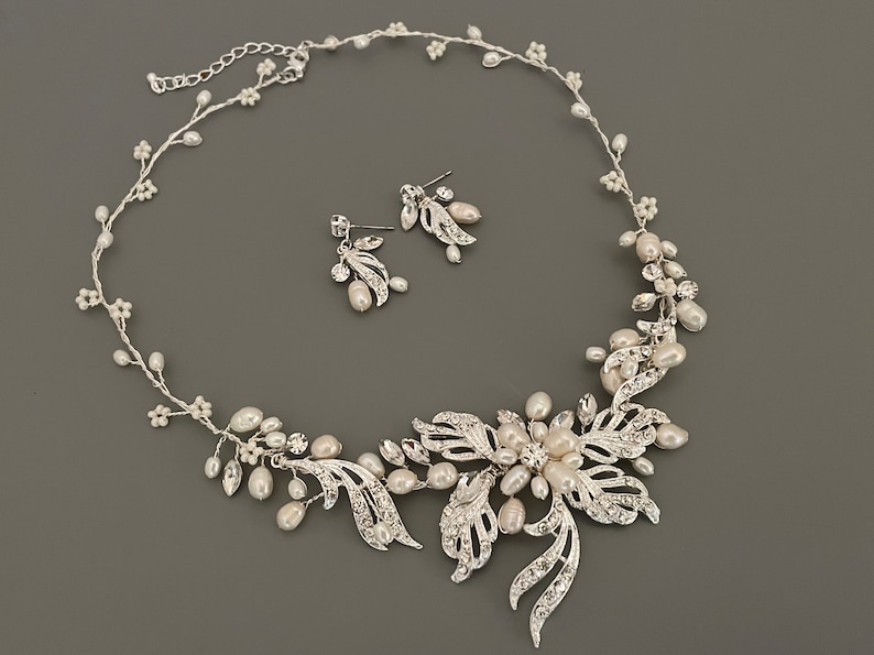 May include: A silver-toned bridal jewelry set featuring a necklace and matching earrings. The necklace has a floral design with faux pearls and clear crystals. The earrings have a similar leaf and pearl design. The set is displayed on a gray background.