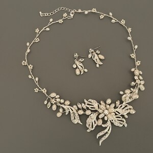 May include: A silver-toned bridal jewelry set featuring a necklace and matching earrings. The necklace has a floral design with faux pearls and clear crystals. The earrings have a similar design. The set is on a gray background.