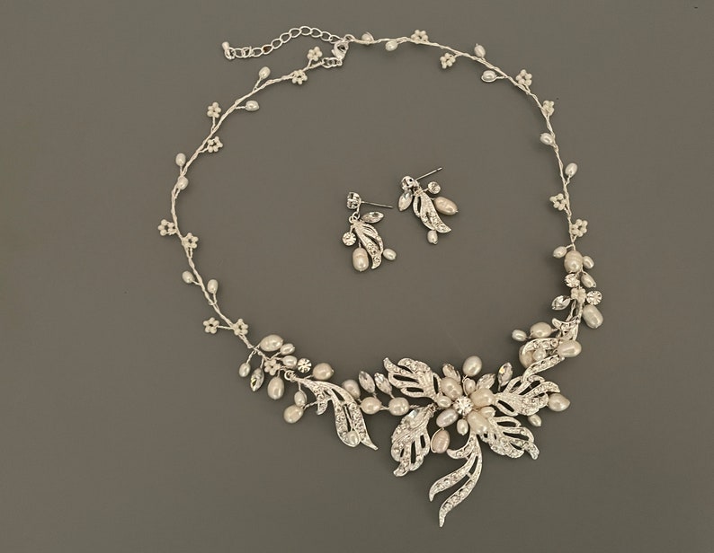 May include: A silver-toned bridal jewelry set featuring a necklace and matching earrings. The necklace has a floral design with faux pearls and clear crystals. The earrings have a similar design. The set is on a gray background.