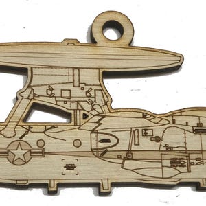 May include: A wooden ornament in the shape of a Navy E-2 Hawkeye aircraft. The aircraft is light brown with etched details, including the text "NAVY" and "E-2". The ornament has a loop at the top for hanging.