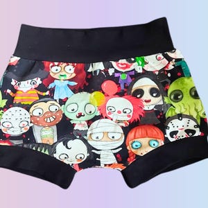 May include: Black shorts with a repeating pattern of cartoon horror characters. The characters include zombies, mummies, clowns, and other monsters. The shorts have a black waistband and leg openings.