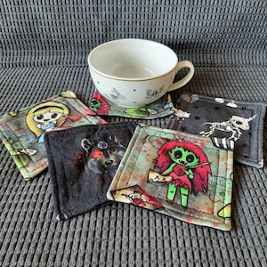 May include: Six colorful fabric coasters with various cartoon characters and designs. The coasters are square and feature a black background with colorful accents. The designs include a girl with a crown, a wolf, a zombie girl, a skeleton, and a butterfly.