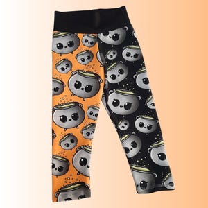 May include: Black and orange leggings with a repeating pattern of cartoon cauldrons. The leggings have a black waistband and a split design, with one leg featuring an orange background and the other a black background.