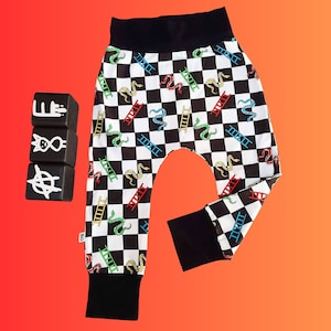 May include: Black and white checkered pants with a pattern of colorful ladders and snakes. The pants have black cuffs and a black waistband. Three black blocks with white symbols are to the left of the pants.