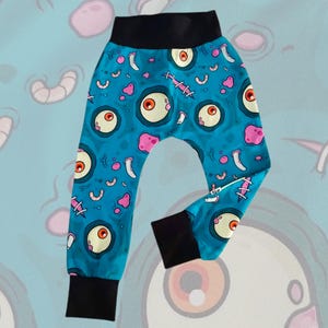 May include: Baby pants with a teal background and a repeating pattern of cartoon eyeballs, pink blobs, and white stitches. The waistband and cuffs are solid black. The pants have a relaxed fit.