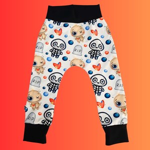 May include: White baby pants with a black waistband and cuffs. The pants feature a repeating pattern of cartoon voodoo dolls, skeleton figures, red hearts, blue circles, and gray tombstones with "R.I.P." text.