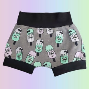May include: Gray baby shorts with a repeating pattern of smiling ice cream popsicles in mint green, pink, and white. The shorts have a black waistband and black trim around the legs.