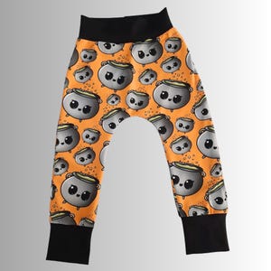 May include: Orange baby pants with a repeating pattern of cartoon cauldrons. The cauldrons are gray with green liquid and have cute faces. The waistband and cuffs are black.