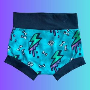 May include: High-waisted shorts with a turquoise background featuring a pattern of white bones and green lightning bolts with purple drips. The waistband and leg openings are black.