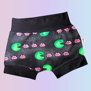 May include: Black baby shorts with a repeating pattern of green Pac-Man-like shapes and pink brain-like figures. The shorts have a black waistband and leg openings. The fabric has a textured, dark gray background.