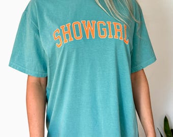 May include: A turquoise t-shirt with the word "SHOWGIRL" in orange block letters across the chest. The shirt has short sleeves and a crew neck. The fabric appears soft and slightly faded.