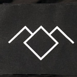 May include: Black fabric patch with a white geometric design. The design features a central diamond shape flanked by two mountain-like triangles. The patch has a textured surface.