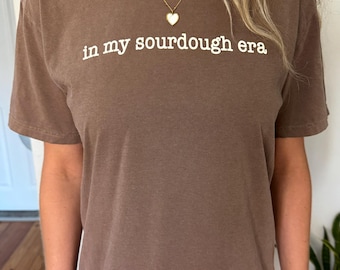 May include: A brown t-shirt with the phrase "in my sourdough era" printed in white. The shirt is a solid color and has short sleeves. A gold necklace with a heart pendant is visible.