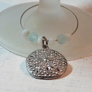 May include: A silver-colored wine charm with a sand dollar pendant. The charm features a wire ring with small, light blue and white beads. The charm is hanging on a frosted glass.