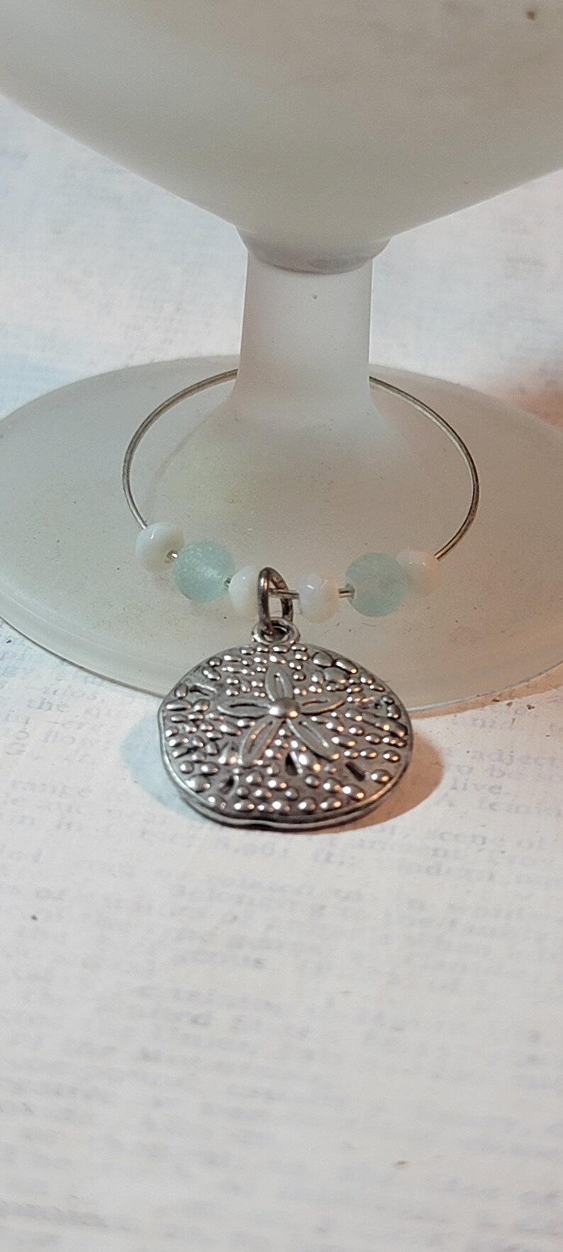 May include: A silver-colored wine charm with a sand dollar pendant. The charm features a wire ring with small, light blue and white beads. The charm is hanging on a frosted glass.