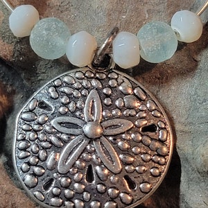 May include: A silver-colored sand dollar pendant with a flower design, strung on a silver wire with white and seafoam green beads. The pendant is detailed with small, textured dots.