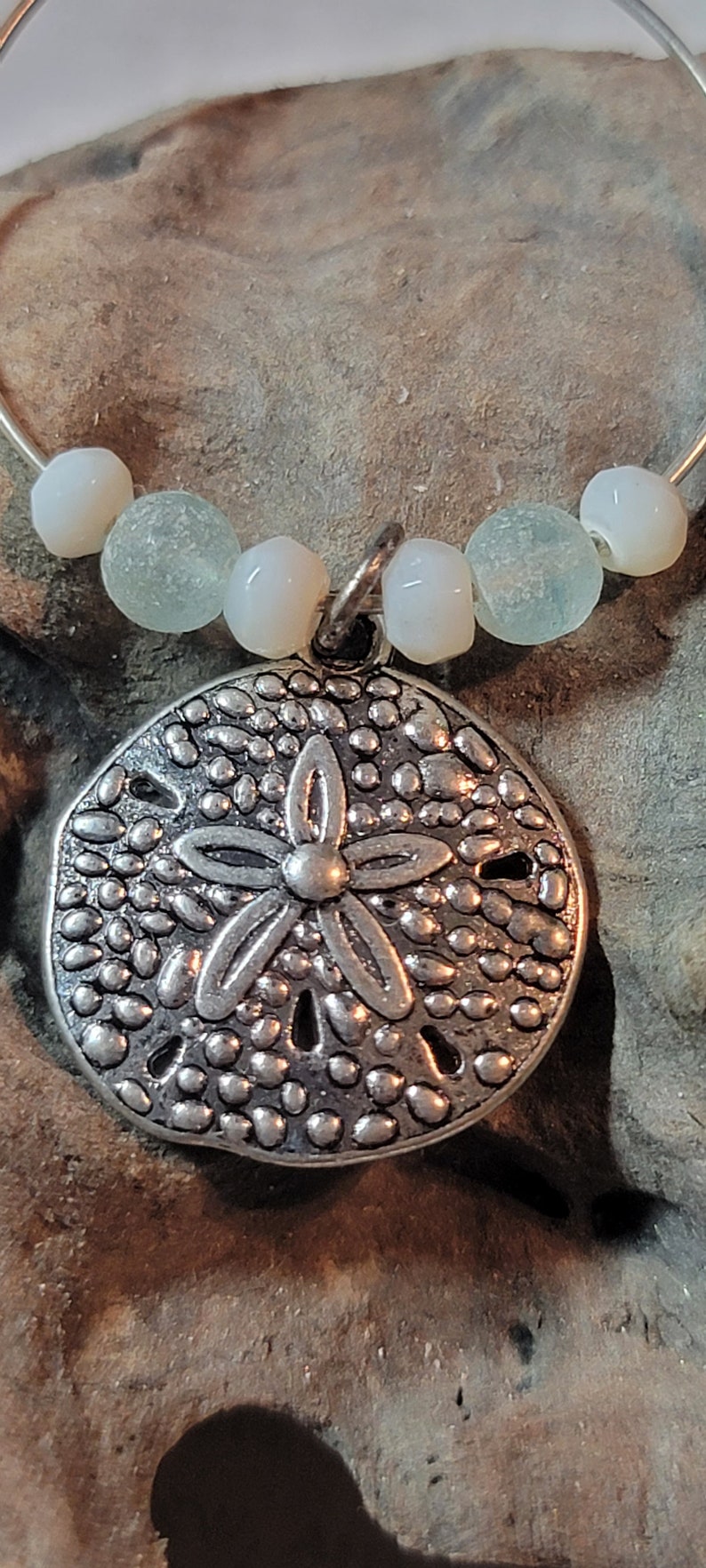 May include: A silver-colored sand dollar pendant with a flower design, strung on a silver wire with white and seafoam green beads. The pendant is detailed with small, textured dots.