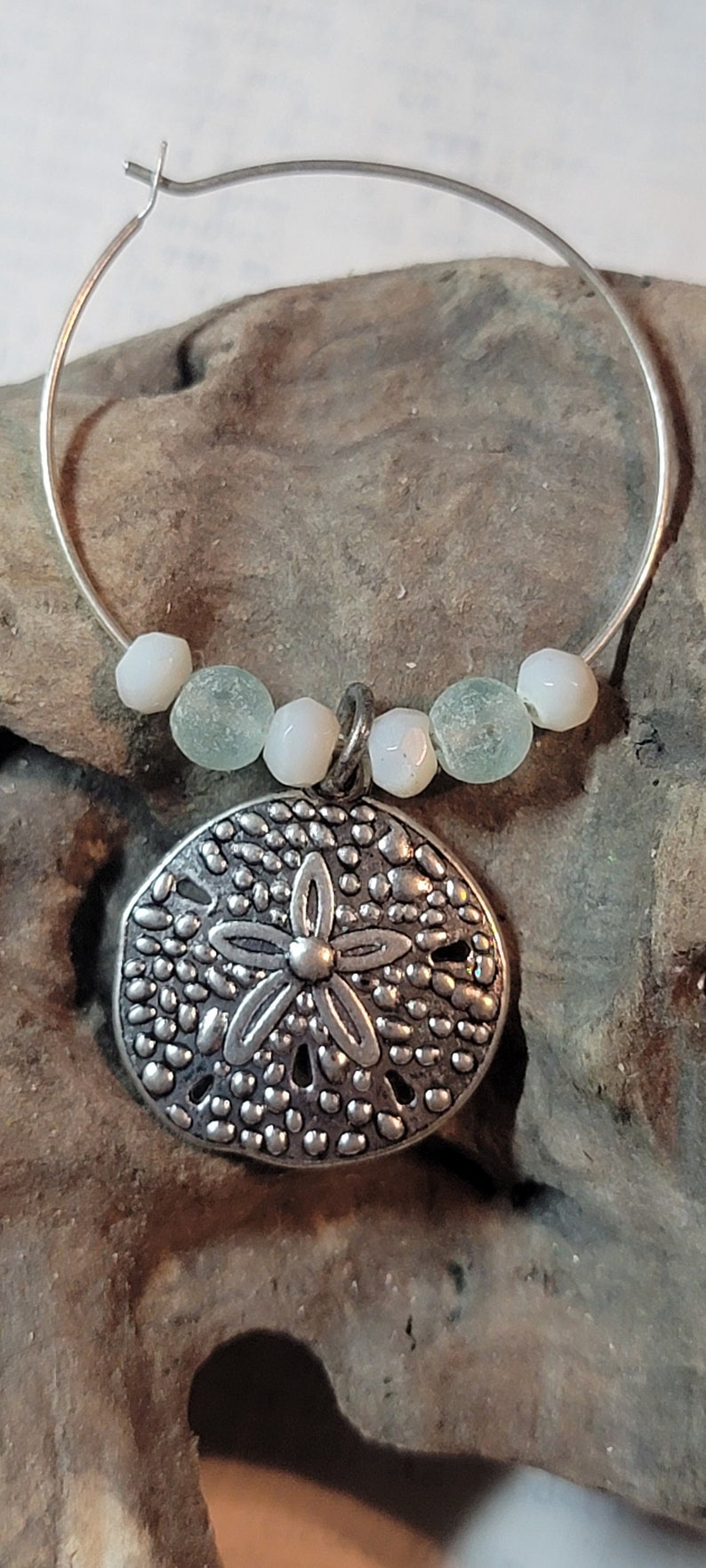 May include: A silver-toned hoop earring with a sand dollar pendant. The pendant is detailed with a floral design and surrounded by small, textured dots. The earring also features small, round, white and seafoam green beads.