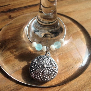 May include: A silver wine charm with a sand dollar pendant, light blue and white beads, and a silver ring. The charm is attached to a wine glass, resting on a wooden surface.