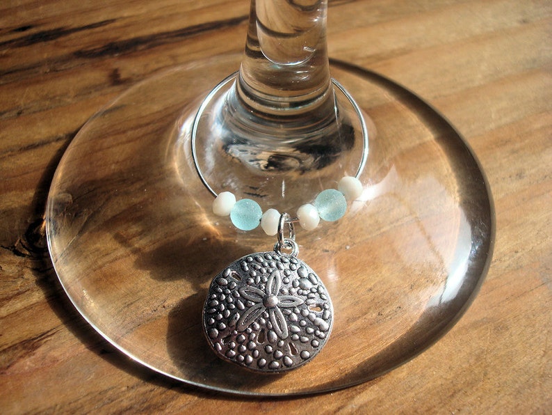May include: A silver wine charm with a sand dollar pendant, light blue and white beads, and a silver ring. The charm is attached to a wine glass, resting on a wooden surface.
