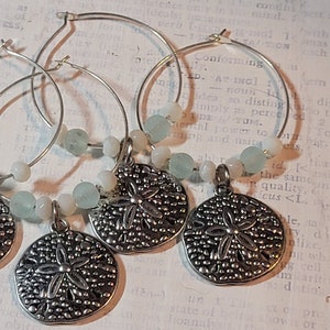 May include: Four silver wine charms with a sand dollar pendant and light blue and white beads. The charms are on silver hoops, perfect for adding a touch of coastal charm to any occasion.