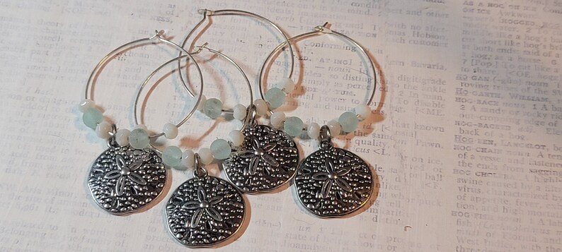 May include: Four silver wine charms with a sand dollar pendant and light blue and white beads. The charms are on silver hoops, perfect for adding a touch of coastal charm to any occasion.