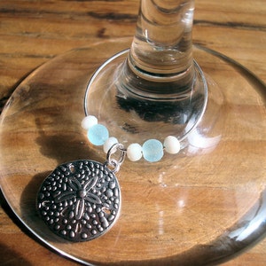 May include: A silver-colored wine charm with a sand dollar pendant and light blue and white beads. The charm is attached to a wine glass, resting on a wooden surface. The sand dollar pendant has a detailed design.