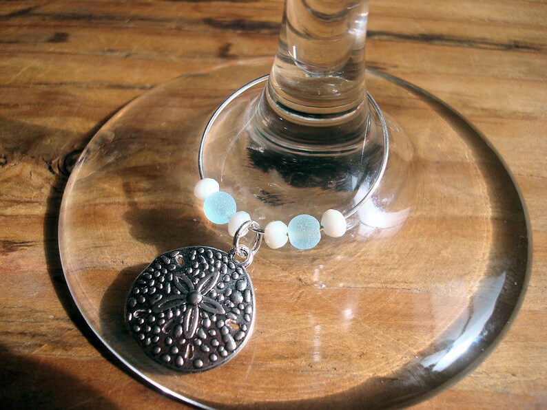 May include: A silver-colored wine charm with a sand dollar pendant and light blue and white beads. The charm is attached to a wine glass, resting on a wooden surface. The sand dollar pendant has a detailed design.