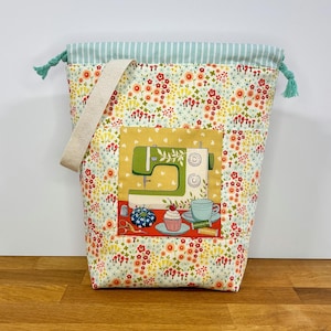 Può includere: Una macchina da cucire verde e bianca con un cupcake e tazze da tè su un piatto rosso, circondata da un motivo floreale in tonalità di giallo, rosso e verde. L'immagine è su una borsa a cordoncino con una parte superiore a righe blu chiaro e bianco.
