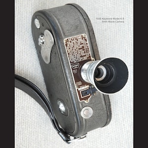 May include: A vintage Keystone Model K-8 8mm movie camera from 1936. The camera is dark gray with a black lens and a leather strap. The text on the camera reads "KEYSTONE" and "8mm Movie Camera".