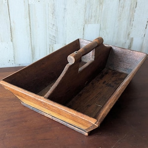 Puede incluir: Un organizador de madera vintage con asa y dos compartimentos interiores. El organizador es de madera marrón oscuro con un acabado natural. El asa es cilíndrica y está unida a los divisores. El organizador es rectangular con lados angulados.