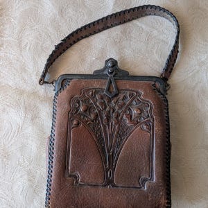May include: Brown leather handbag with a decorative floral design and a stitched handle. The bag has a metal clasp and trim. The leather has a worn appearance, suggesting it is vintage or antique.