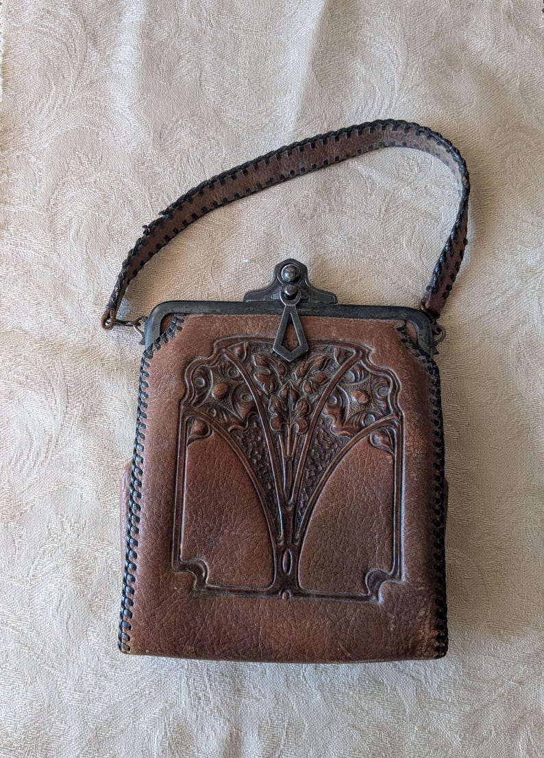 May include: Brown leather handbag with a decorative floral design and a stitched handle. The bag has a metal clasp and trim. The leather has a worn appearance, suggesting it is vintage or antique.