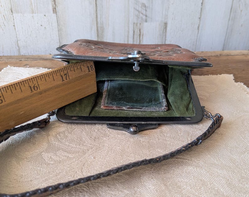 May include: Vintage brown leather purse with a metal clasp and chain strap. The purse is open, revealing a green interior and a small mirror. A wooden ruler is placed next to the purse, showing measurements in inches.