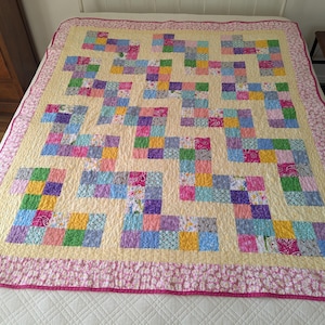 May include: A patchwork quilt featuring a zigzag pattern of multi-colored fabric squares in shades of pink, blue, yellow, green, and purple, set against a pale yellow background with a wide pink floral border and a thin pink edge.