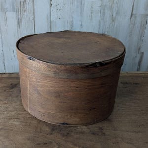 Könnte beinhalten: Eine runde, dunkelbraune Aufbewahrungsbox aus Holz mit passendem Deckel. Der Behälter hat einen zylindrischen Körper aus dünnem, gebogenem Holz mit sichtbarer Maserung und einer verwitterten, strukturierten Oberfläche an Deckel und Gehäuse.
