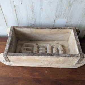 May include: A rectangular, weathered wooden box with metal banding. The interior features a raised, stylized design. The box has rounded ends and sits on a dark wooden surface, with a light-colored wooden wall in the background.