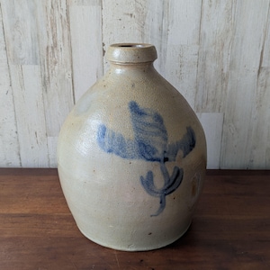 May include: A stoneware jug with a rounded body and narrow neck. The jug is a light beige color with a blue floral design on the front. The jug is sitting on a wooden surface.