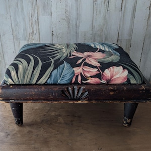 May include: A rectangular footstool with a dark brown wooden frame featuring a carved shell detail at the center. The upholstered top displays a black fabric base with a printed pattern of large green palm leaves and pink hibiscus flowers.