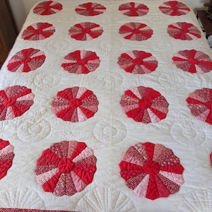 May include: A handmade quilt with a white base, adorned with a repeating pattern of red and pink pinwheel motifs. The quilt is crafted from diverse fabrics and has a quilted texture, representing traditional craftsmanship.