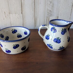 Könnte beinhalten: Ein Keramik-Schüssel- und Krug-Set. Die cremefarbenen Stücke sind mit blauen Blaubeer-Designs und grünen Blättern verziert. Beide Teile haben einen blauen Rand. Der Krug hat einen Griff und einen Ausguss. Die Schüssel ist oval.