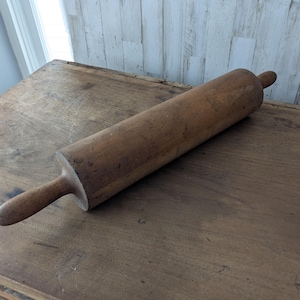 May include: A cylindrical wooden rolling pin with a smooth, weathered brown finish and two tapered handles on either end. The surface of the rolling pin shows natural wood grain patterns, minor surface scratches, and signs of age and use.