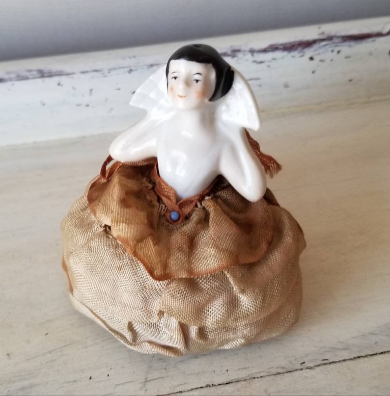 May include: A vintage porcelain pincushion featuring a seated doll with black hair, a white collar, and a brown and gold dress. The doll has a painted face with a smile.