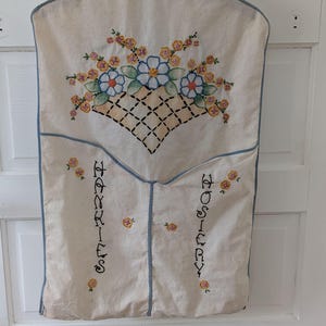 May include: A cream-colored fabric hanging organizer with blue trim, featuring embroidered blue and yellow flowers in a basket design. The two lower pockets are labeled with black embroidered text reading HANKIES and HOSIERY respectively.