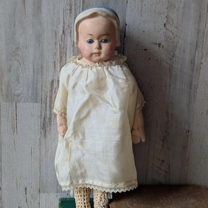 Puede incluir: Una muñeca antigua con rostro pálido, ojos azules y rasgos pintados, que lleva un gorro azul claro y un vestido color crema de manga corta con ribete de encaje en el cuello y el dobladillo. La muñeca tiene manos de tela y pies de ganchillo.