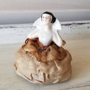 May include: A porcelain doll with black hair and a white collar sits on a beige fabric pincushion. The doll is wearing a brown and gold dress with a blue jewel accent.