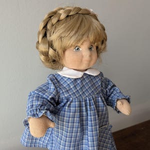 Op de afbeelding: Een handgemaakte pop met blond haar in een gevlochten kroon, die een blauw-wit geruit jurkje draagt met een witte kraag.