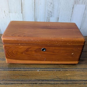 Può includere: Una scatola di legno rettangolare con coperchio incernierato e un piccolo buco della serratura rotondo. La scatola è realizzata in legno dai toni caldi, con motivi di venature visibili e un aspetto leggermente usurato. La scatola è appoggiata su una superficie di legno.