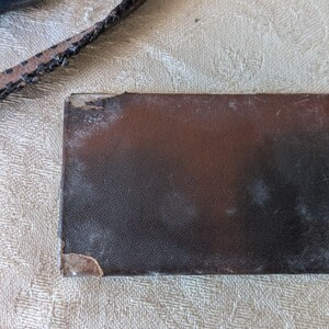 May include: A rectangular, dark brown leather wallet with a textured surface. The wallet shows signs of wear, with a worn corner and some light discoloration. A portion of a leather strap is visible in the upper left corner.