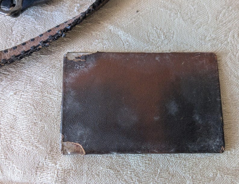 May include: A rectangular, dark brown leather wallet with a textured surface. The wallet shows signs of wear, with a worn corner and some light discoloration. A portion of a leather strap is visible in the upper left corner.